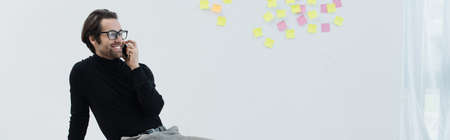 pleased man in eyeglasses talking on mobile phone near sticky notes on gray wall, bannerの写真素材