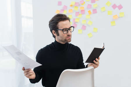 man in eyeglasses and black turtleneck working with notebook and document near sticky notes on gray...