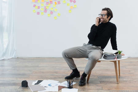happy man talking on mobile phone while sitting on coffee table near papers on floor