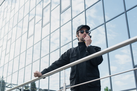 low angle view of guard in black uniform and sunglasses talking on walkie-talkie near building with...