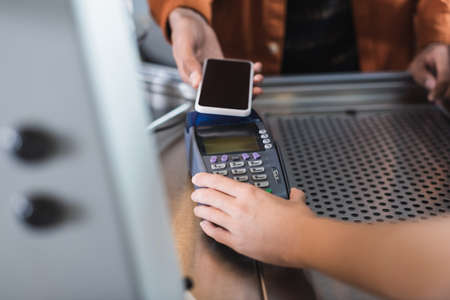 Cropped view of barista holding payment terminal near african american customer with cellphone in...