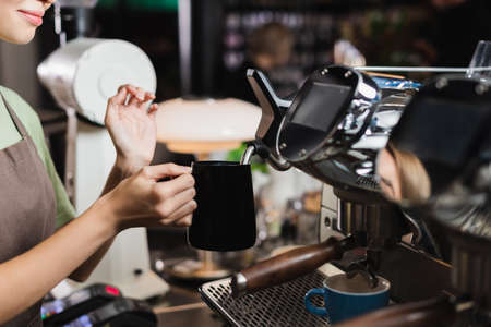 Cropped view of young barista holding jug near steam wand of coffee machine in cafeの写真素材