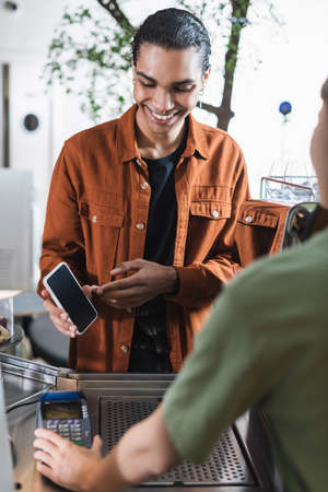 Smiling african american customer pointing with smartphone near blurred barista in cafe