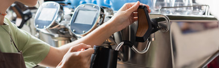 Cropped view of barista holding jug near steam wand of coffee machine in cafeteria, banner