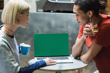 Side view of multiethnic freelancers holding coffee and using laptop with green screen in cafe
