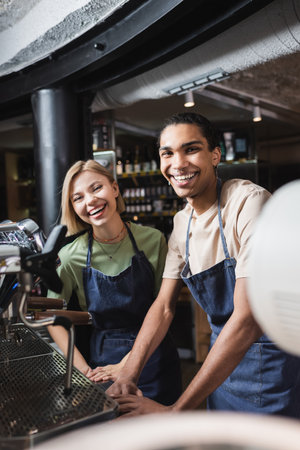 Cheerful interracial baristas looking at camera near coffee machine in cafe