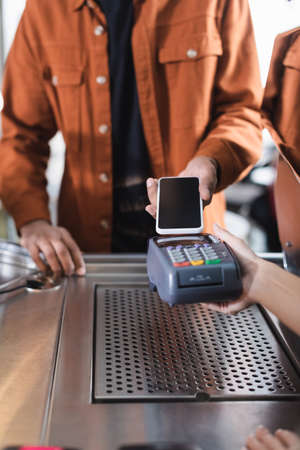 Cropped view of barista holding payment terminal near african american customer with smartphone in...