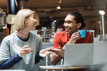 Smiling interracial freelancers talking and holding coffee near devices in cafe