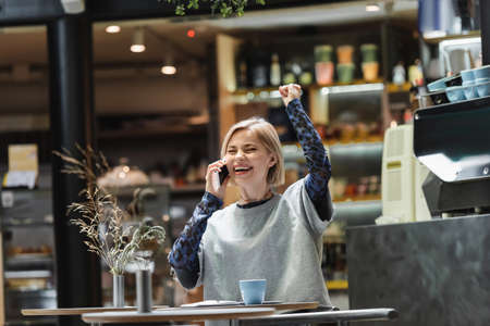 Excited blonde woman talking on smartphone near coffee and notebook in cafeの写真素材
