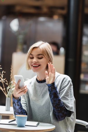 Positive woman having video call on smartphone near coffee and notebook in cafe