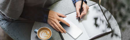 Cropped view of woman writing on notebook near smartphone and cup of coffee in cafe, banner
