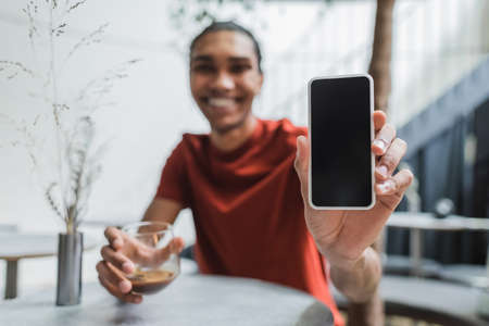 Smartphone with blank screen in hand of blurred african american man with coffee in cafeの写真素材