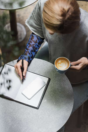 Overhead view of blonde woman writing on notebook near cellphone and holding cup in cafe