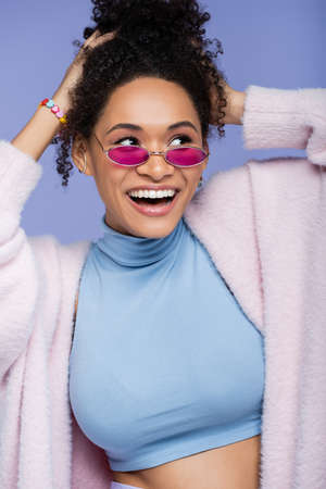 cheerful african american woman in pink sunglasses adjusting curly hair isolated on violetの写真素材