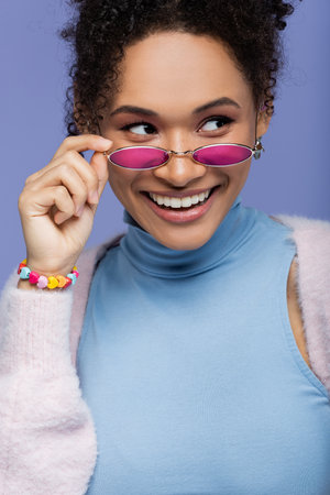 positive african american woman adjusting trendy sunglasses isolated on violet