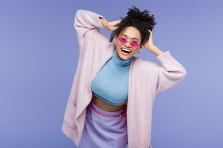 happy african american woman in stylish sunglasses adjusting curly hair isolated on violet