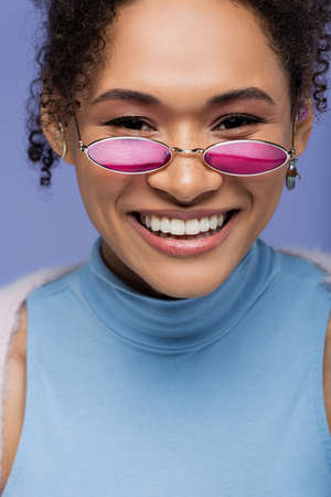 close up of pleased african american woman in pink sunglasses smiling isolated on violetの写真素材