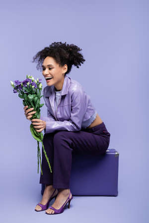cheerful african american woman sitting on suitcase and holding flowers on purpleの写真素材