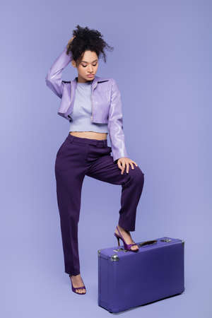 full length of stylish african american woman standing near suitcase on purpleの写真素材