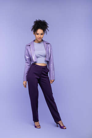 full length of curly african american woman in leather jacket and trousers posing on purpleの写真素材