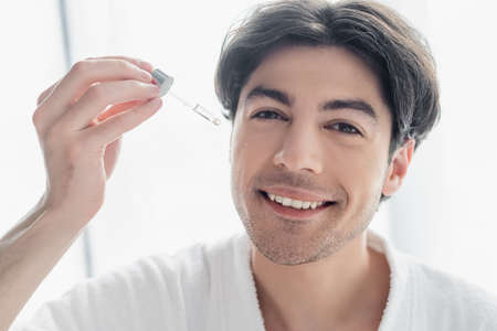 happy man looking at camera while applying cosmetic serum in bathroom