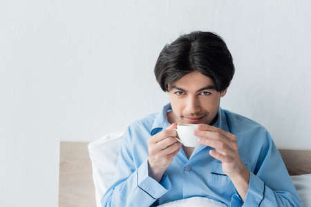brunette man smiling at camera while drinking coffee in bedroomの写真素材