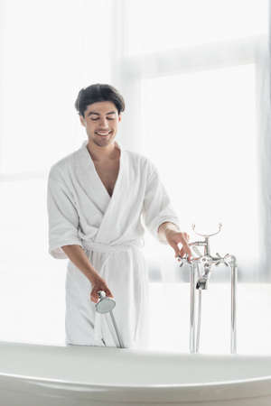 smiling man in white bathrobe holding shower head while opening faucet in bathroom