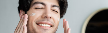 close up view of smiling man with closed eyes applying face cream, banner