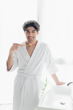 joyful man in white bathrobe looking at camera while holding toothbrush with toothpaste in bathroomの写真素材