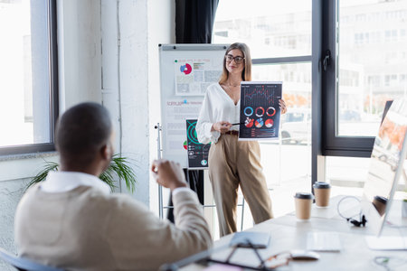 smiling businesswoman in glasses showing infographics to african american man in office