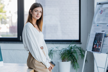 cheerful young businesswoman holding paper cup and leaning on desk in officeの写真素材