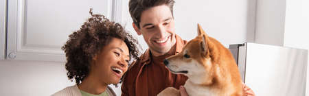 funny shiba inu dog near smiling interracial couple in kitchen, bannerの写真素材