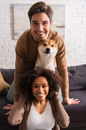 Positive interracial couple looking at camera near shiba inu dog on couch at home