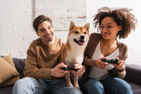 KYIV, UKRAINE - DECEMBER 22, 2021: Multiethnic couple looking at shiba inu and playing video game in...