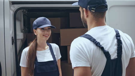 Positive delivery woman in uniform looking at colleague near cardboard boxes in car outdoorsの写真素材