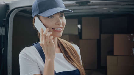 Positive delivery woman talking on mobile phone near blurred packages in car outdoorsの写真素材