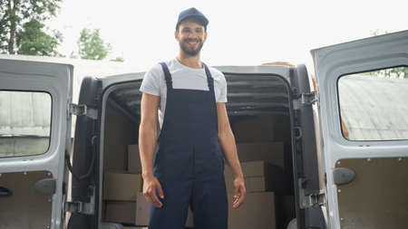 Smiling courier in overalls standing near cardboard packages in car outdoorsの写真素材