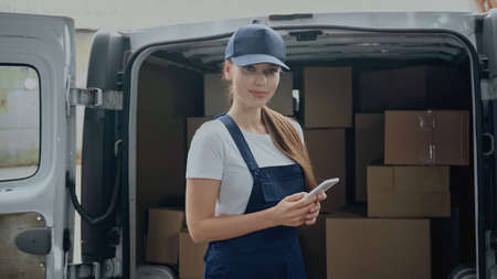 Delivery woman in overalls holding smartphone and looking at camera near cardboard boxes in car...