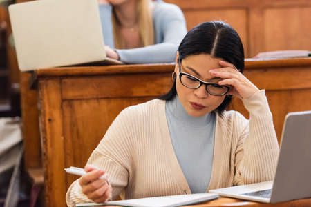 Asian student looking at notebook near laptop during lecture in university