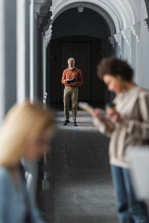 Mature teacher looking at notebook near blurred interracial students in corridor of universityの写真素材
