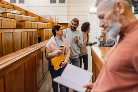 Smiling multiethnic students with coffee to go talking near friends and blurred teacher at universityの写真素材