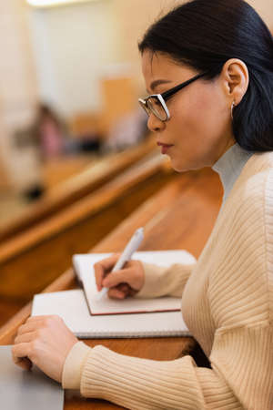 Side view of asian student writing on blurred notebook near laptop in university