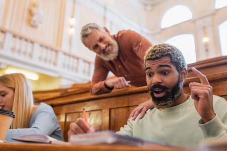 Excited african american student writing on notebook near blurred teacher in universityの写真素材
