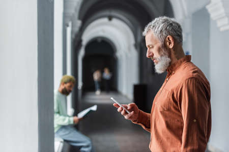 Side view of mature teacher using smartphone in corridor of university
