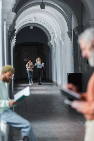 Interracial students with coffee and laptop walking on university corridor