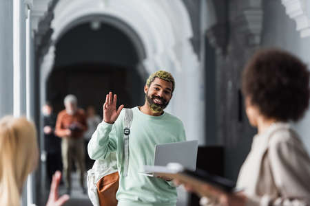 African american student with laptop waving hand at blurred friends in university corridorの写真素材