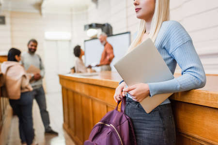 Blonde student holding backpack and laptop near blurred friends in universityの写真素材
