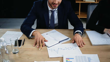 cropped view of businessman in formal wear near empty notebook and graphs on desk
