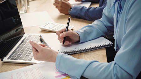 cropped view of businesswoman writing in notebook near laptop with blank screen