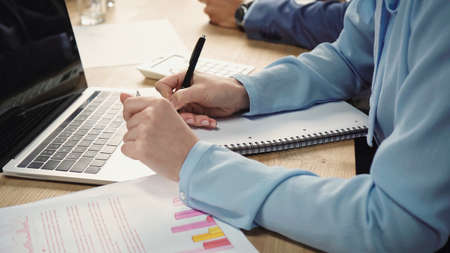 cropped view of businesswoman writing in notebook near graphs and laptop with blank screen